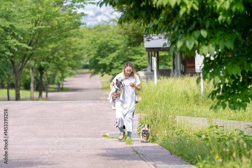 On a summer morning in a park with blooming hydrangeas, a Japanese woman in her 30s walks two French Bulldogs. Surrounded by flowers and trees, they stroll along quiet paths under soft morning light.