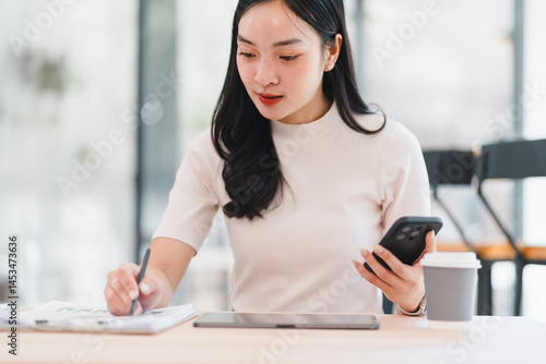 Free Download Young Asian woman working at desk with smartphone, tablet, and coffee cup, writing notes on paper in bright modern office, focused and productive atmosphere - GFXLoad