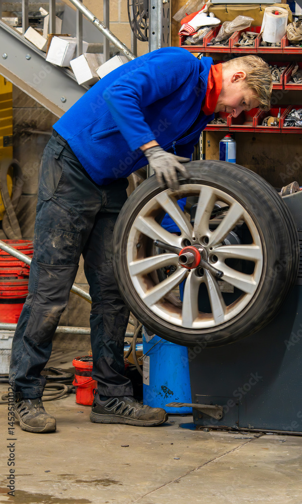 An auto technician balancing wheel and tire assembly in a professional garage workshop. Mechanic in blue jacket handling alloy rim on tire balancing machine during seasonal tire replacement service An auto technician balancing wheel and tire assembly in a professional garage workshop. Mechanic in blue jacket handling alloy rim on tire balancing machine during seasonal tire replacement service