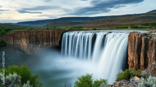 Majestic wide-angle image of a powerful waterfall flowing over rocky cliffs surrounded by lush green trees under a cloudy sky du daytime, captu natural beauty and serene landscape scenery