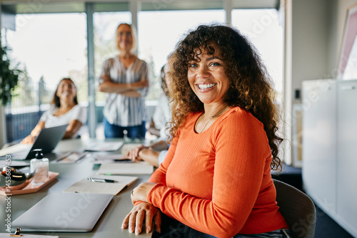 Confident woman with curly hair and orange top smiling in modern office setting. Documents, laptop, and snacks are spread out on the meeting table. Bright and positive atmosphere