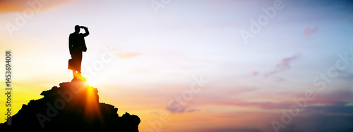 Slika na platnu Businessman standing on mountain peak at sunrise looking forward with hand over eyes, symbolizing vision and leadership in business strategy concept