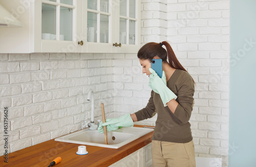 Bild auf Leinwand Focused young woman wearing rubber gloves using cup plunger to unclog sink in kitchen while talking on phone