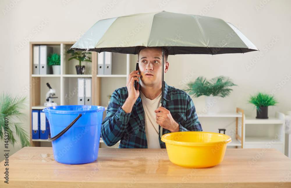 Worried man sitting at desk under umbrella, making phone call to repair and maintenance service. Office worker dealing with plumbing or roof problem and leaking water dripping into bucket and basin.