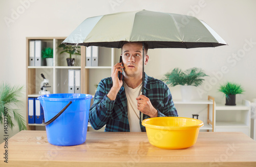 Obraz na płótnie Worried man sitting at desk under umbrella, making phone call to repair and maintenance service. Office worker dealing with plumbing or roof problem and leaking water dripping into bucket and basin. w salonie