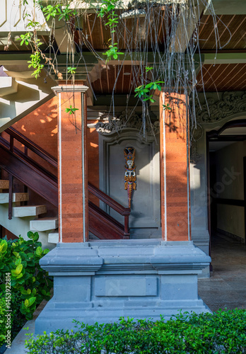 Traditional Balinese entryway or home entrance with red columns, ornate woodwork, hanging plants, and a decorative deity figure mounted on the wall.