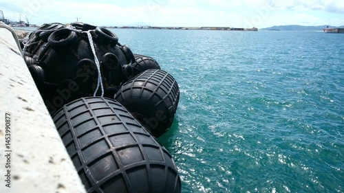 Wide angle view of quayside equipment at Gibraltar. Floating pneumatic rubber fenders secured to moorings are moved about by the strong winds and waves. Above the bay is a blue sky with white clouds.
