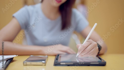 A woman is writing on a tablet with a pen. She is sitting at a table with a cell phone and a keyboard