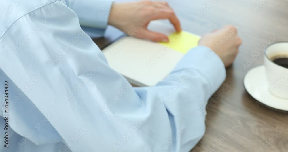 Woman putting sticky note onto notebook and writing memo at wooden table, closeup Woman putting sticky note onto notebook and writing memo at wooden table, closeup