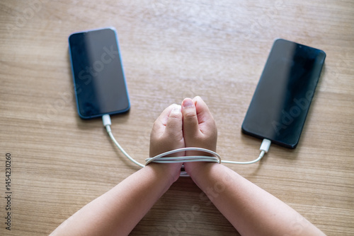 Quadro em tela Child's Hands tied with charging cables between two smartphones on wooden table, symbolizing smartphone addiction and dependency in modern digital life
