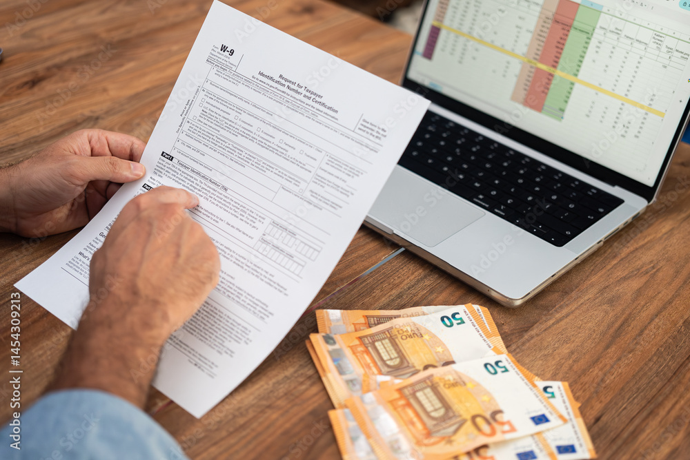 Obraz premium Property landlord inspects W-9 tax form to supply TIN for reporting rental income and real estate transactions on desk with euro bills and laptop nearby
