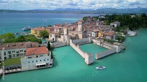 Drone flyaway from Scaliger Castle in Sirmione, Italy, revealing turquoise waters of Lake Garda, colorful houses, and historic walls on a sunny spring day.