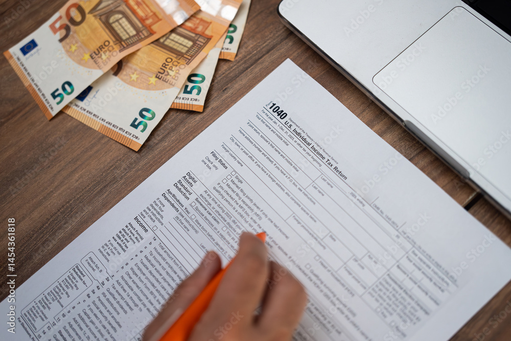 Obraz premium Male freelancer carefully examines strict requirements for filling Individual Income Tax Return form sitting at workplace with euro banknotes and laptop