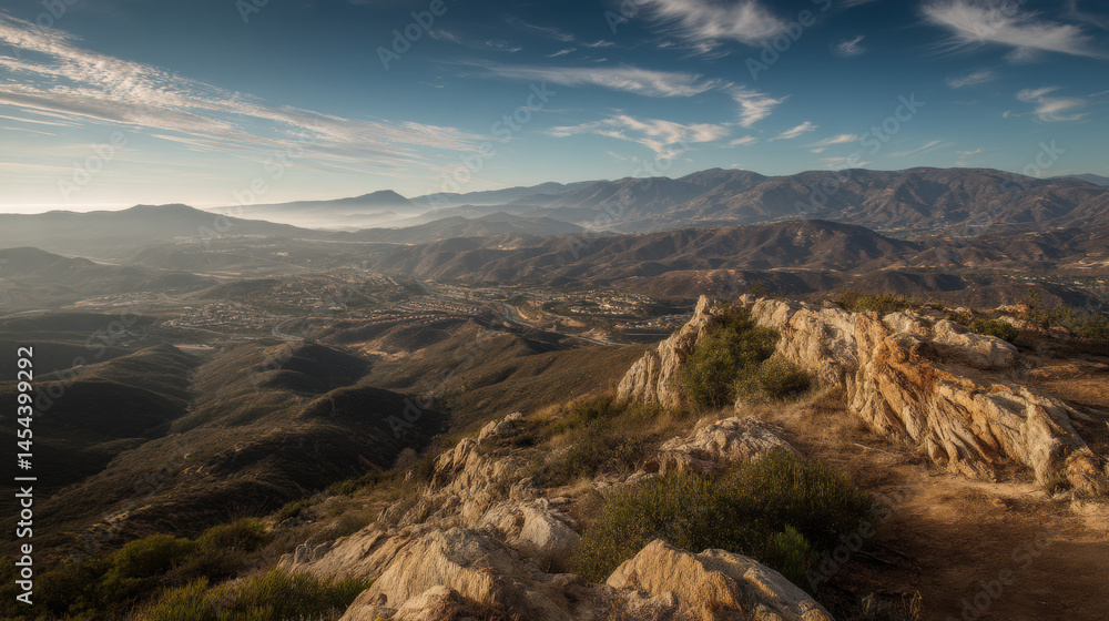 Obraz premium An expansive vista unfolds with rocky outcroppings overlooking a valley shrouded in mist, mountains rising in the distance under a bright blue sky at sunrise.