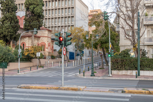 An urban street in Athens, Greece, lined with trees and modern buildings. A traffic light glows red, while a blue road sign directs towards Syntagma Square. A hill with buildings rises in the backgrou