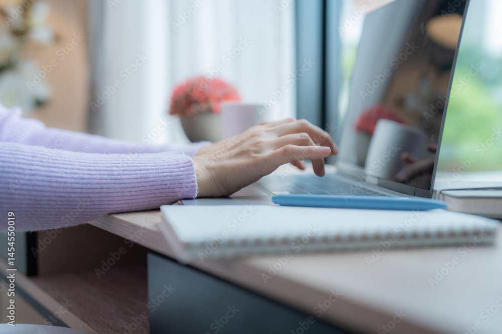 Fototapeta premium Woman's hands are typing on a laptop keyboard placed on a wooden desk with a notebook and pen next to it, suggesting a home office environment
