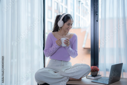 Free Download Happy freelancer woman wearing headphones, drinking coffee and listening to music while sitting on windowsill with laptop nearby, enjoying her break time at home - GFXLoad