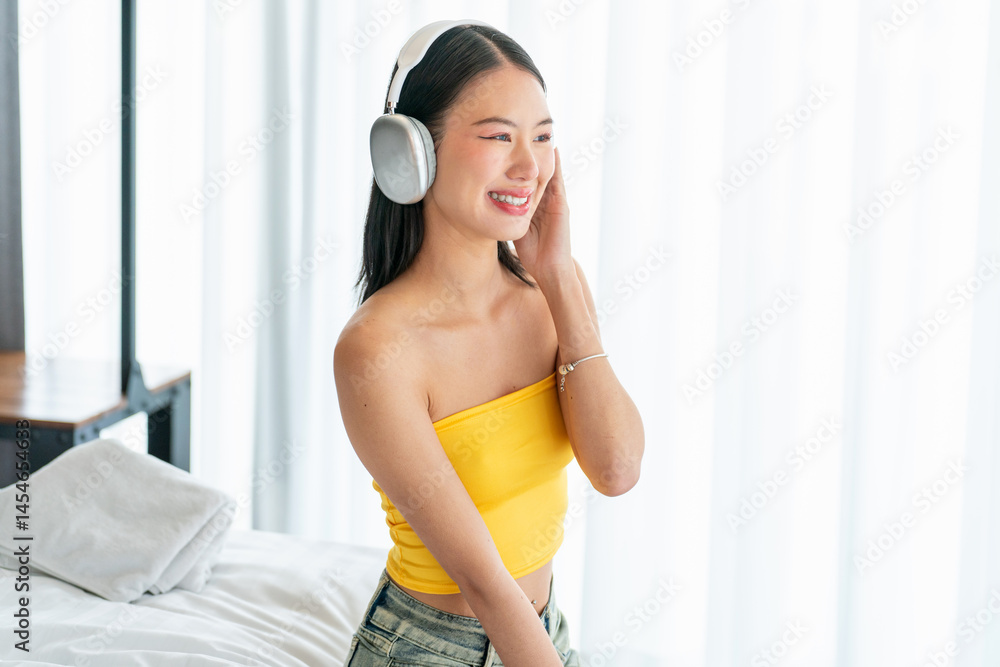 Happy Asian Woman Enjoying Music on Headphones While Sitting on Bed in Bright Room Happy Asian Woman Enjoying Music on Headphones While Sitting on Bed in Bright Room