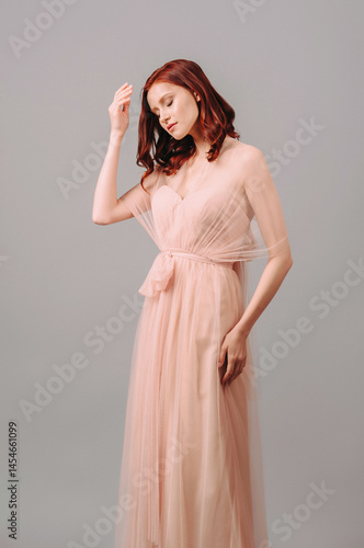 Elegant bridesmaid’s two-layer maxi gown. Gorgeous young sexy lady with curly hairstyle. Pink chiffon evening dress with bow detail. Seductive ginger woman with sheer cape in studio