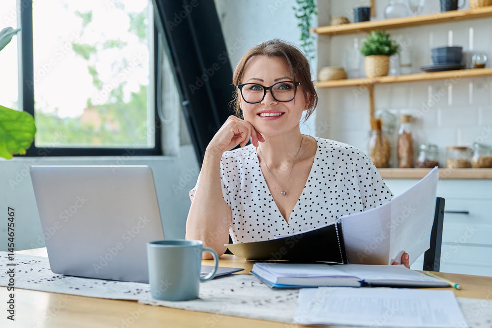 Middle aged smiling woman working with laptop, looking at camera, sitting at home