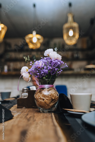 Photo of decor, design, glass vase with paper inside, with artificial flowers of lilac and white peonies on wooden table in restaurant.