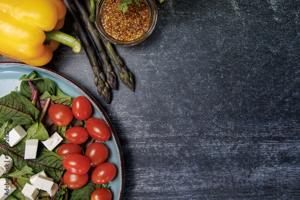 healthy food on a wooden table, top view.Salad with cherry tomatoes, cubes of white cheese and greens on a dark surface. next to bell peppers, asparagus, mustard seeds, top view, space for text. healthy food on a wooden table, top view.Salad with cherry tomatoes, cubes of white cheese and greens on a dark surface. next to bell peppers, asparagus, mustard seeds, top view, space for text.