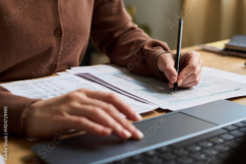 Closeup of hands of unrecognizable young woman calculating expenses using laptop, while sitting at desk and managing budget