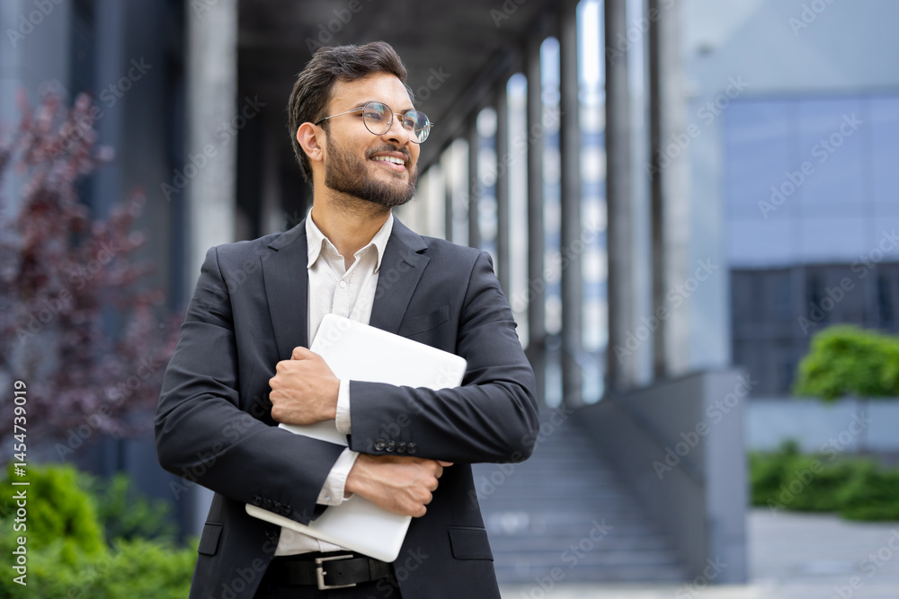 A confident businessman with glasses stands outdoors holding a laptop, smiling brightly near a modern building.