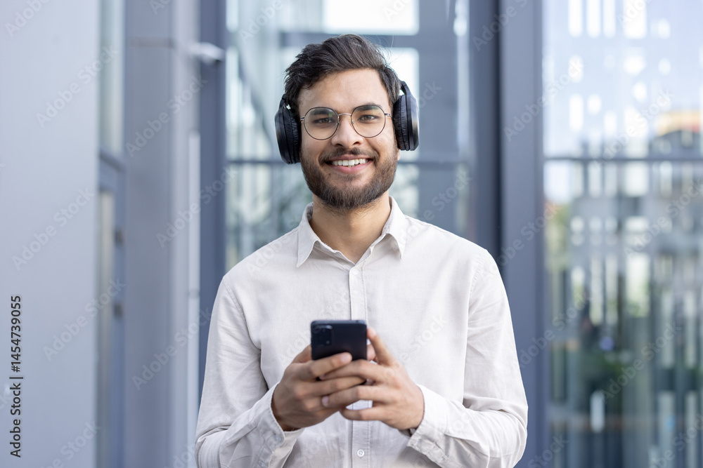 A smiling man wearing headphones and glasses holds his smartphone, possibly listening to music in an office setting.