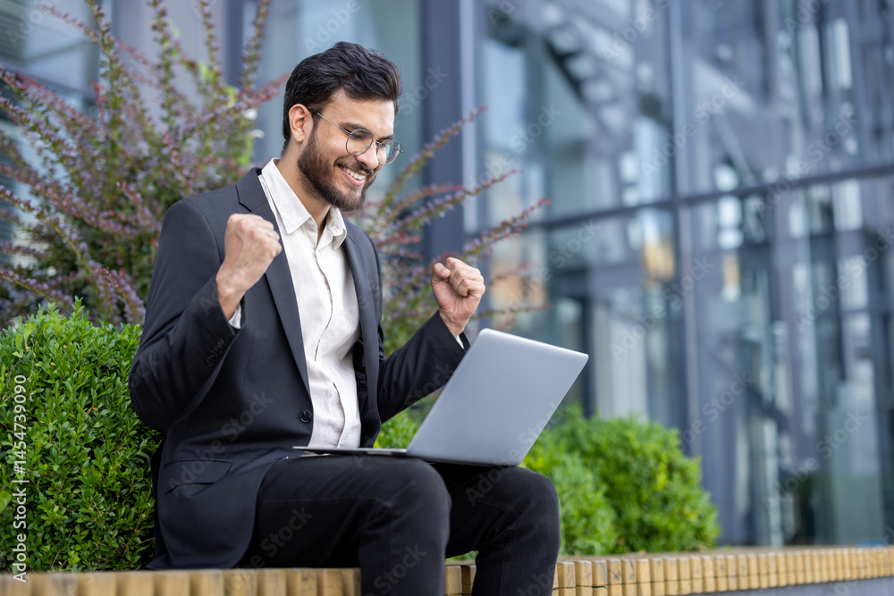 A successful businessman celebrates after reviewing positive results on his laptop outside, smiling with excitement.