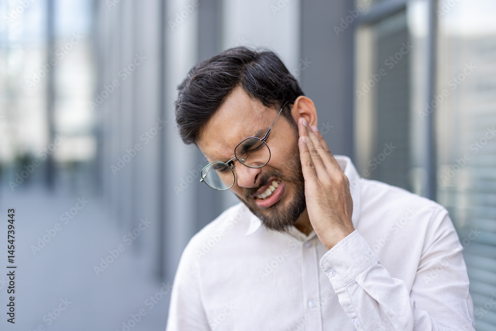 A man in a white shirt is seen with a pained expression, touching his neck and ear outside a building.