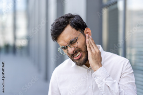 Obraz na płótnie A man in a white shirt is seen with a pained expression, touching his neck and ear outside a building. w salonie
