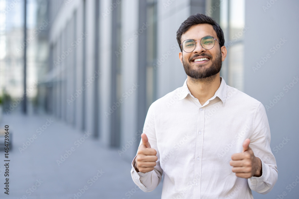 A cheerful businessman shows a thumbs-up gesture, standing outdoors with a bright smile, celebrating success or approval.