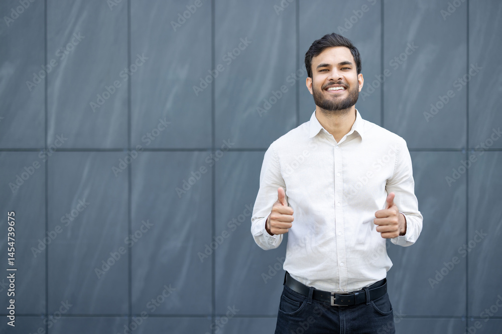 A smiling man with dark hair and a beard shows thumbs up, dressed in a white shirt. The background is a gray textured wall.