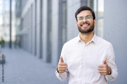Obraz na płótnie A cheerful businessman shows a thumbs-up gesture, standing outdoors with a bright smile, celebrating success or approval. w salonie