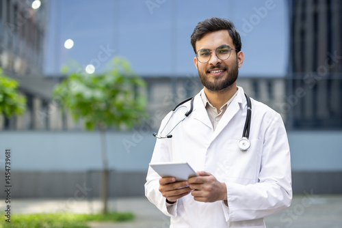 Obraz na płótnie A smiling doctor with glasses holds a tablet while standing outdoors with a stethoscope around his neck. w salonie
