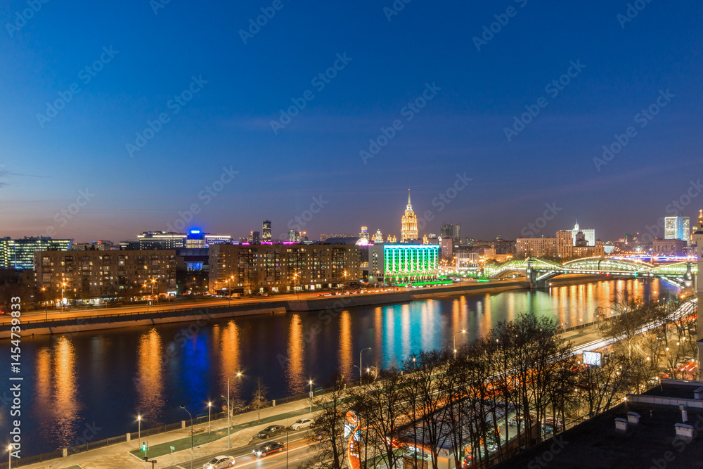 Obraz premium The skyline of Moscow glows at night with colorful lights reflecting in the river. Busy streets and bridges show the city's vibrant nightlife and urban energy.