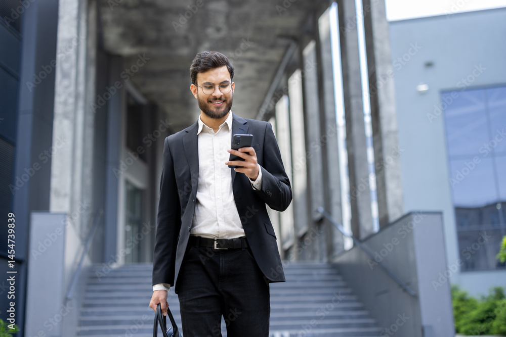 A young businessman in a suit looks at his phone while walking outdoors near a modern building, holding a briefcase.