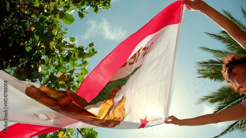 A woman holding the State California flag against a strong wind, with a background of clear blue sky and tropical trees.