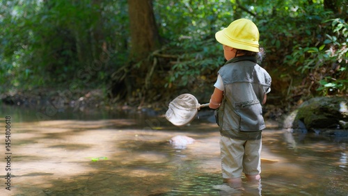 A young Asian child wearing a yellow hat and vest explores a forest stream with a fishing net, surrounded by lush greenery on a Sunny Day, ready to Discovering the wonders of nature