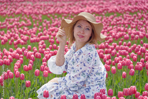 Na zdjęciu widoczna jest osoba siedząca w rozległym polu pełnym czerwonych tulipanów. Ma na sobie kapelusz i kwiecistą sukienkę, co dodaje sielankowego uroku. W tle można dostrzec zieloną roślinność i