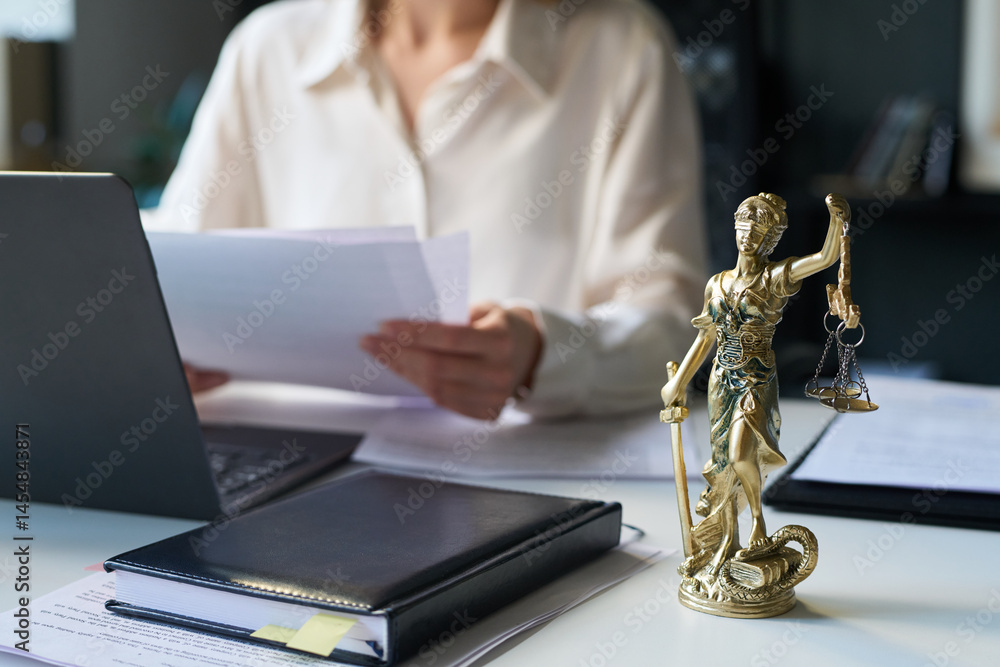 © pressmaster - Bronze Lady Justice statuette on desk of attorney in law firm, holding scales and sword, and female lawyer working with documents in blurred background