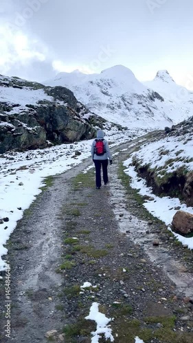 POV of a woman with a red backpack walking along a snow covered mountain path, surrounded by snowy peaks, rocks, and a flowing stream in a serene winter landscape
