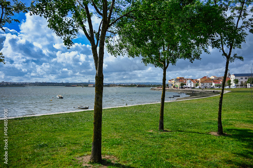 Trees and grass along the Tejo River in Seixal, Portugal, with boats, traditional houses, and a cloudy sky on a peaceful afternoon.