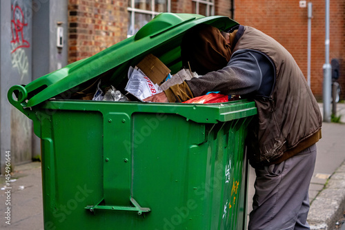 Homeless person is rummaging through a green dumpster on a city street, searching for items.