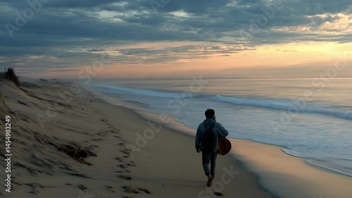 Solitary figure strolls sandy beach at dawn carrying guitar, leaving footprints in the wet sand, watching ocean waves.