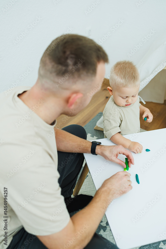 Dad teaches his 1.6 year old son to sculpt from plasticine. The toddler boy sits at the children's table with his dad and plays entertaining games. The child sculpts figures from plasticine