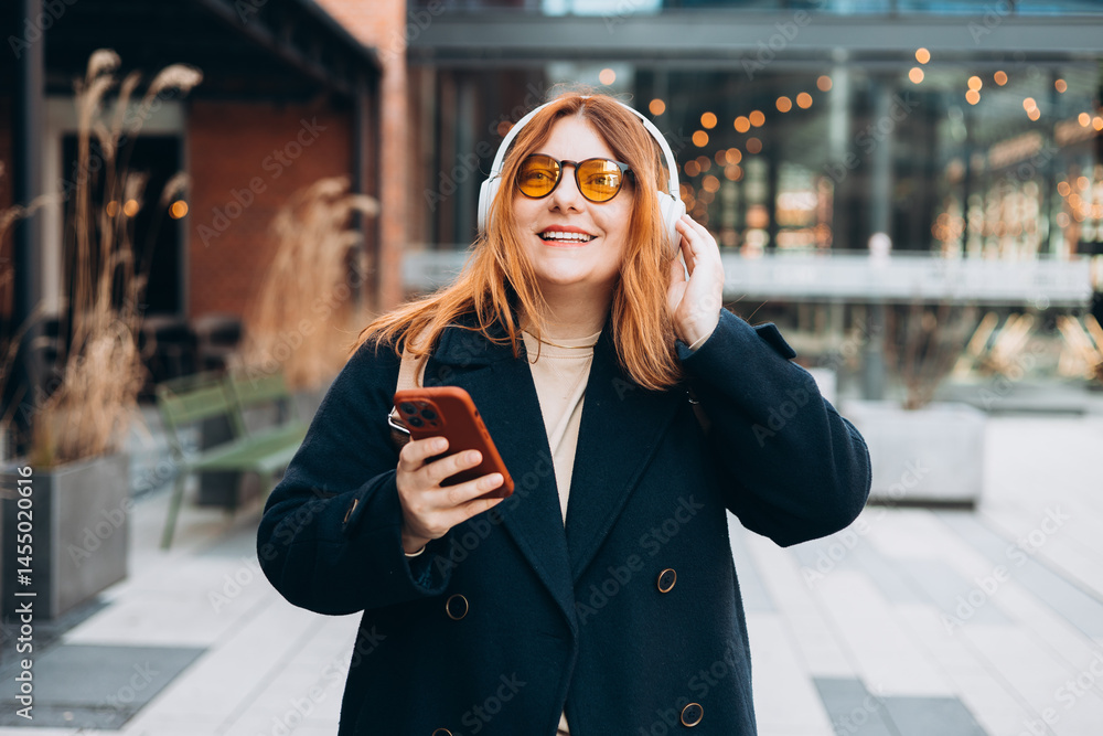 30s millennial Caucasian lady student in wireless headphones with phone listen music, enjoy audio lesson in university campus, outdoor. Woman holding smartphone on urban background.