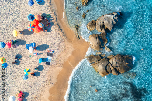 Fototapeta Naklejka Na Ścianę i Meble -  Aerial view of colorful umbrellas, sandy beach and blue sea with waves and rocks at sunset. Summer in Sardinia island, Italy, Tropical seascape. Top drone view of clear water, relaxing people, beach