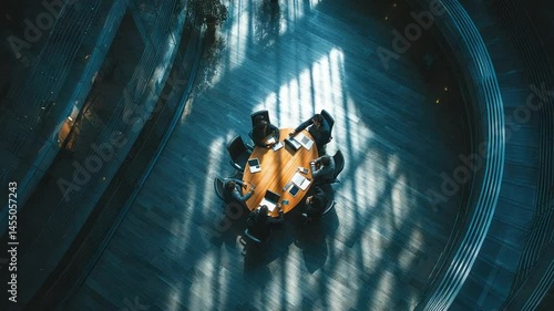 High-angle view of a business team gathered around a circular table in a modern, sophisticated office space. Sunlight streams through the structure, casting dramatic shadows